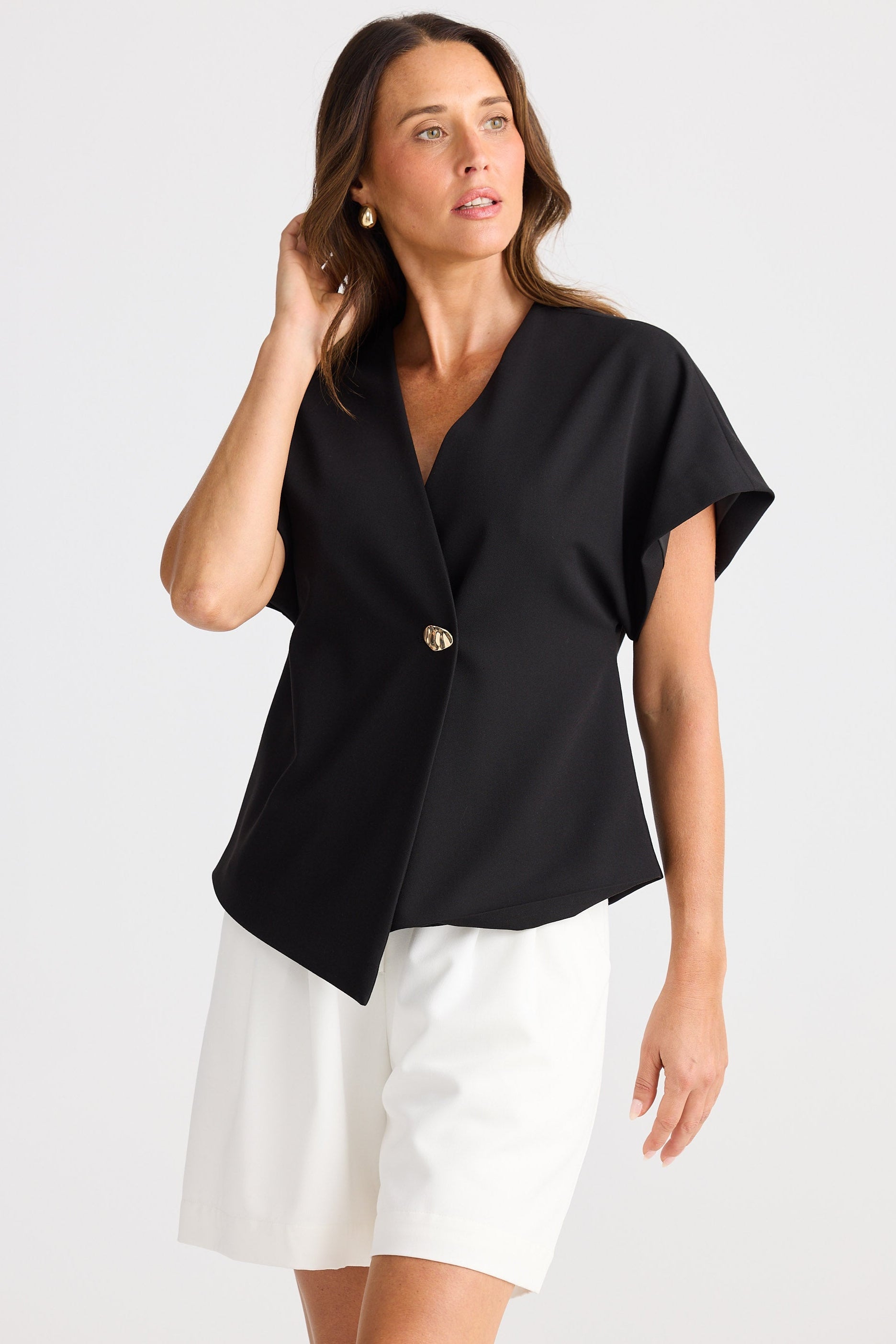 Woman wearing a black top with a gold button and white shorts on a light gray background