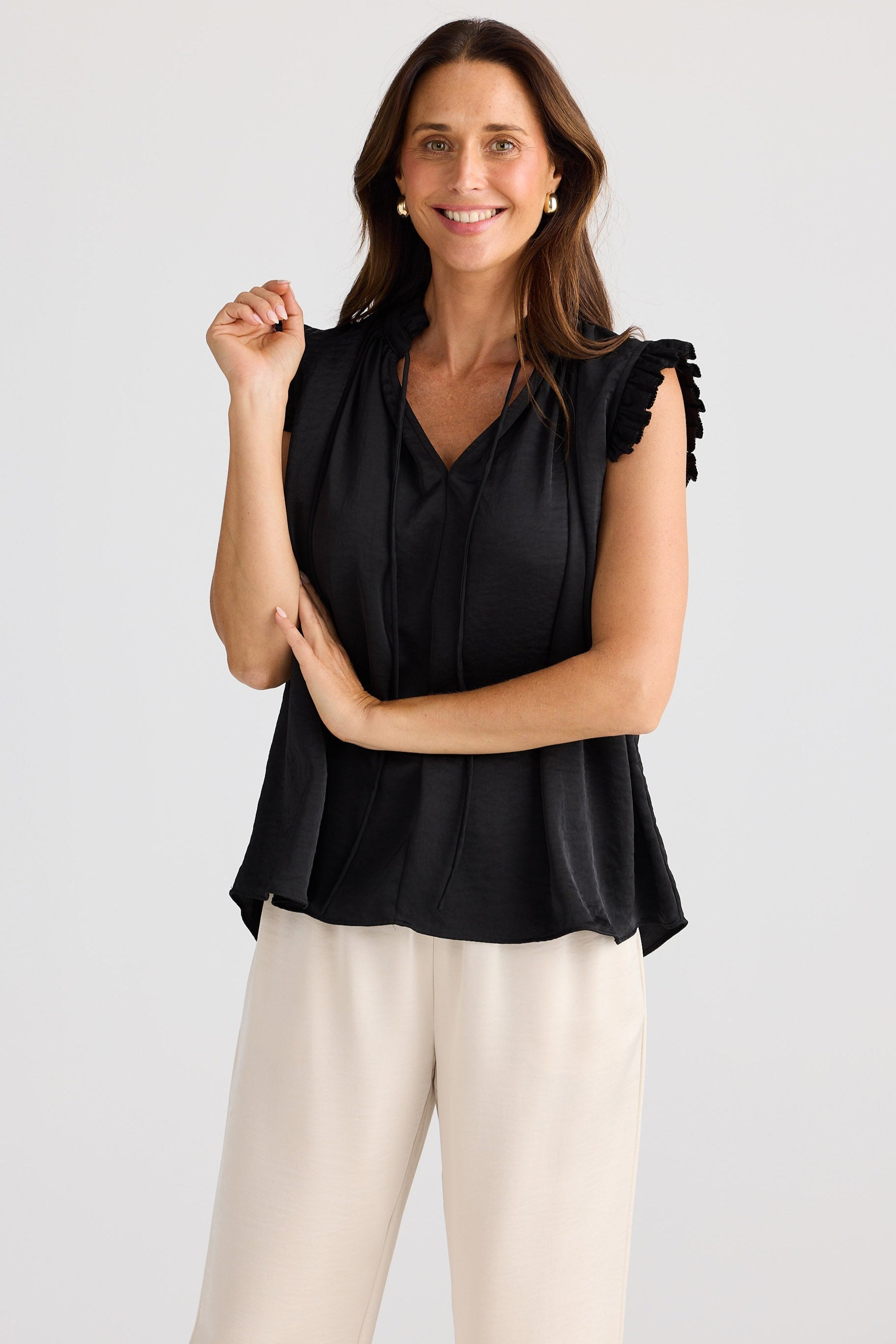 Woman wearing a black sleeveless top with ruffled shoulders and beige pants on a white background